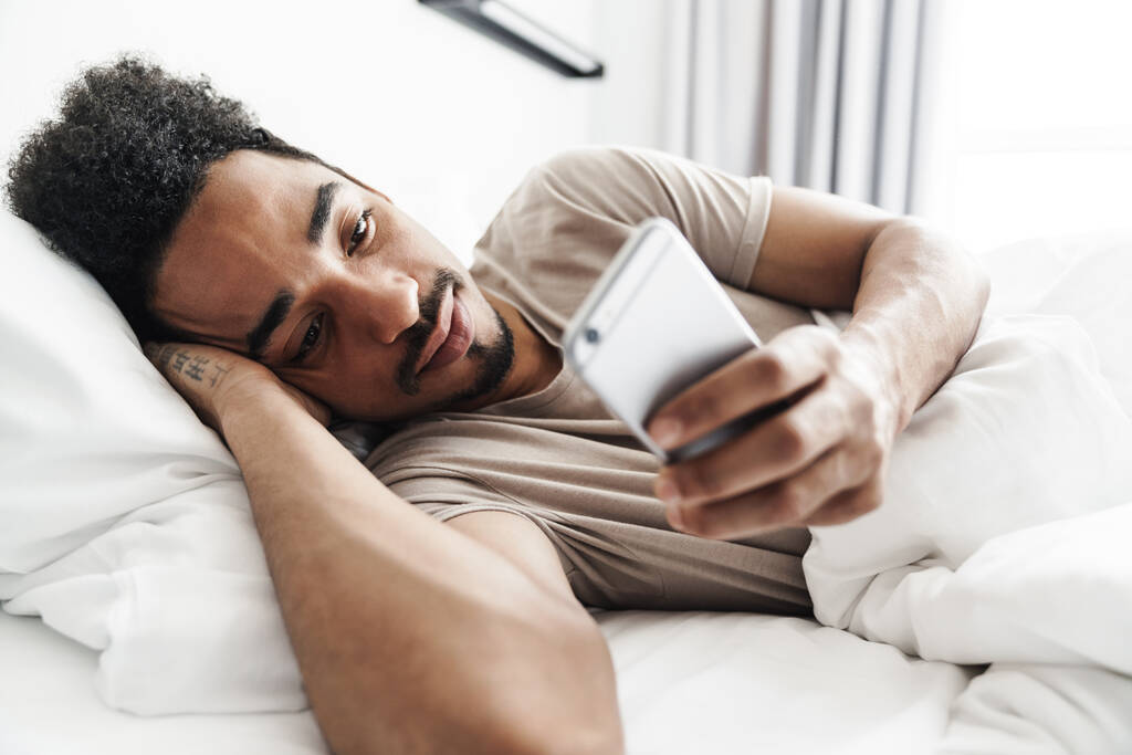 Φωτογραφία του ευχαριστημένου Αφροαμερικανού με μουστάκι που χρησιμοποιεί κινητό τηλέφωνο σε λευκό κρεβάτι στο φωτεινό δωμάτιο - Φωτογραφία, εικόνα
