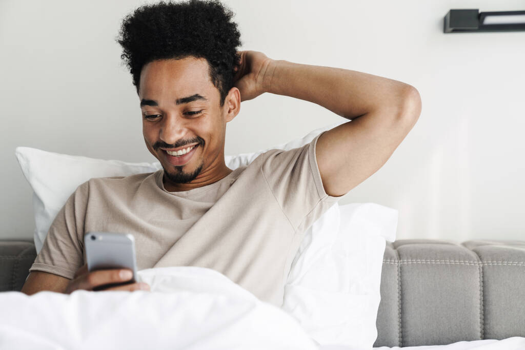 Photo d'un homme afro-américain heureux avec une moustache utilisant un téléphone portable et souriant au lit dans une pièce lumineuse - Photo, image