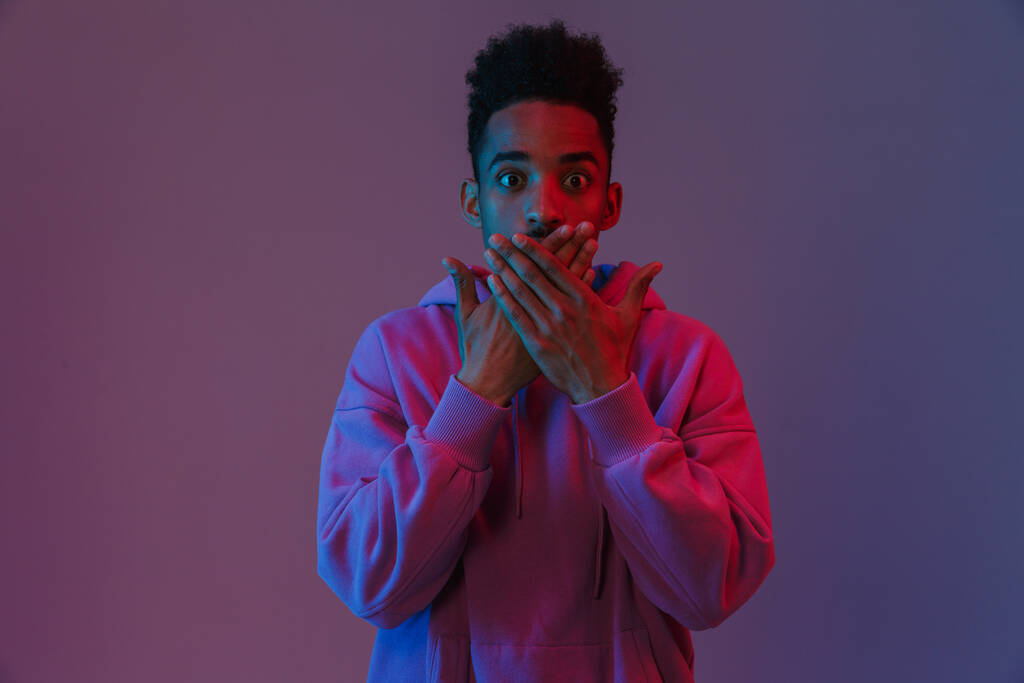 Ritratto di uomo afroamericano sorpreso in felpa con cappuccio colorato che si posa davanti alla macchina fotografica con le mani sulla bocca isolate su sfondo viola - Foto, immagini