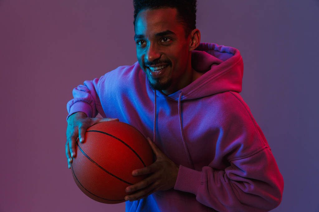 Retrato de homem americano africano feliz com capuz colorido batendo na câmera com basquete isolado sobre fundo violeta
 - Foto, Imagem