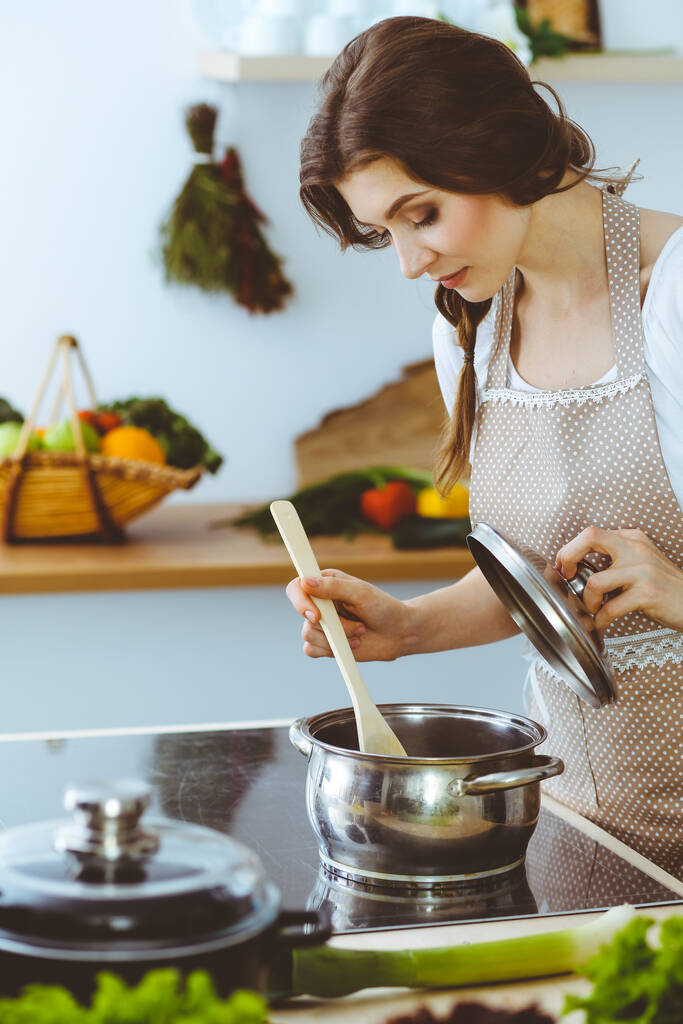 Genç esmer kadın mutfakta çorba pişiriyor. Ev hanımı elinde tahta kaşık tutuyor. Gıda ve sağlık konsepti - Fotoğraf, Görsel