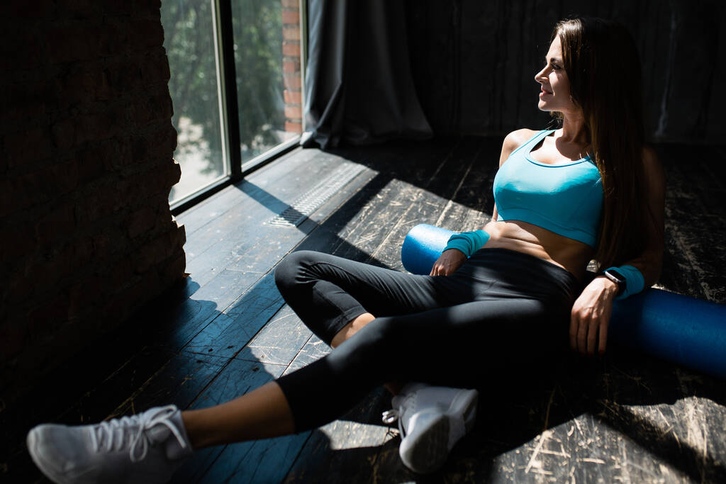 Mujer entrenador de fitness en una parte superior azul y polainas negras apoya sus manos en el suelo y hace ejercicio la fascia lumbar con un cilindro de espuma
 - Foto, imagen