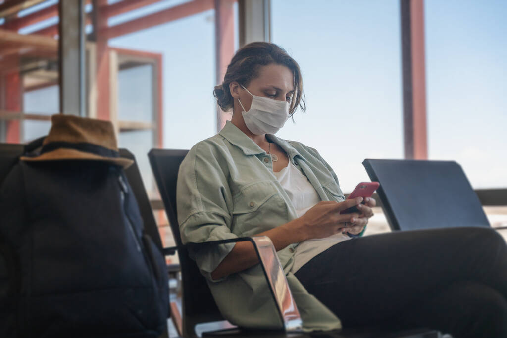 Jonge vrouw reiziger met een beschermend medisch masker met een smartphone in haar handen wacht op haar vlucht op de luchthaven, een nieuwe normale levensstijl. - Foto, afbeelding