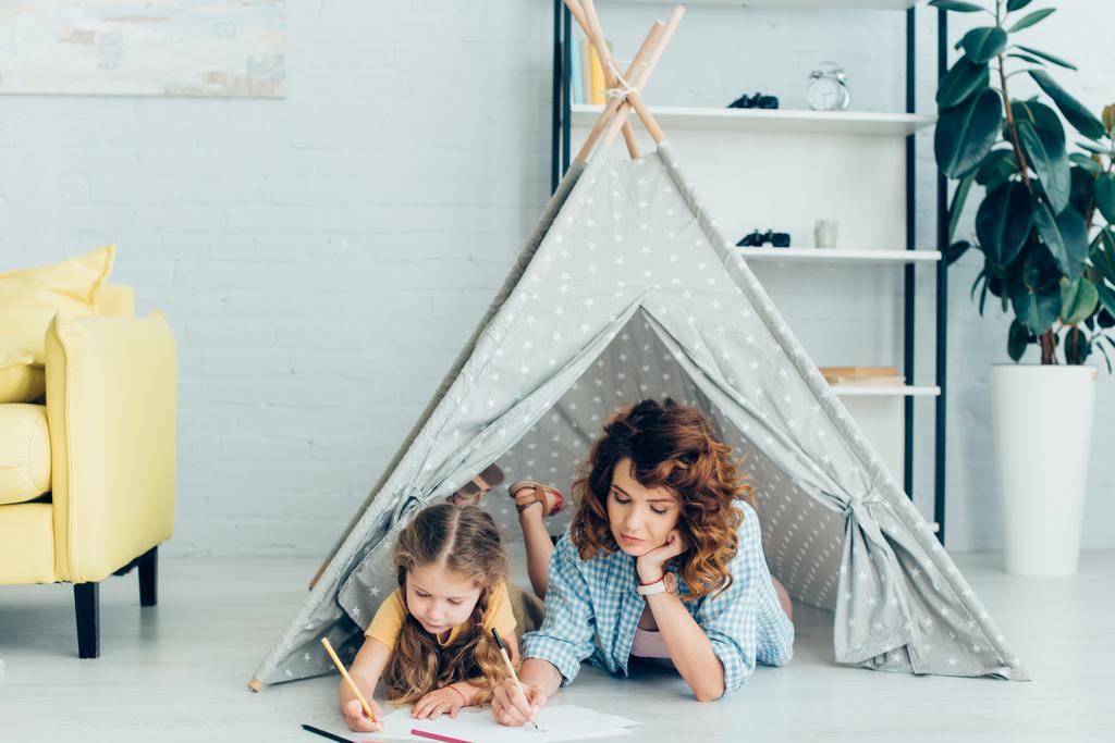 young nanny and cute kid drawing together while lying in toy wigwam - Photo, Image
