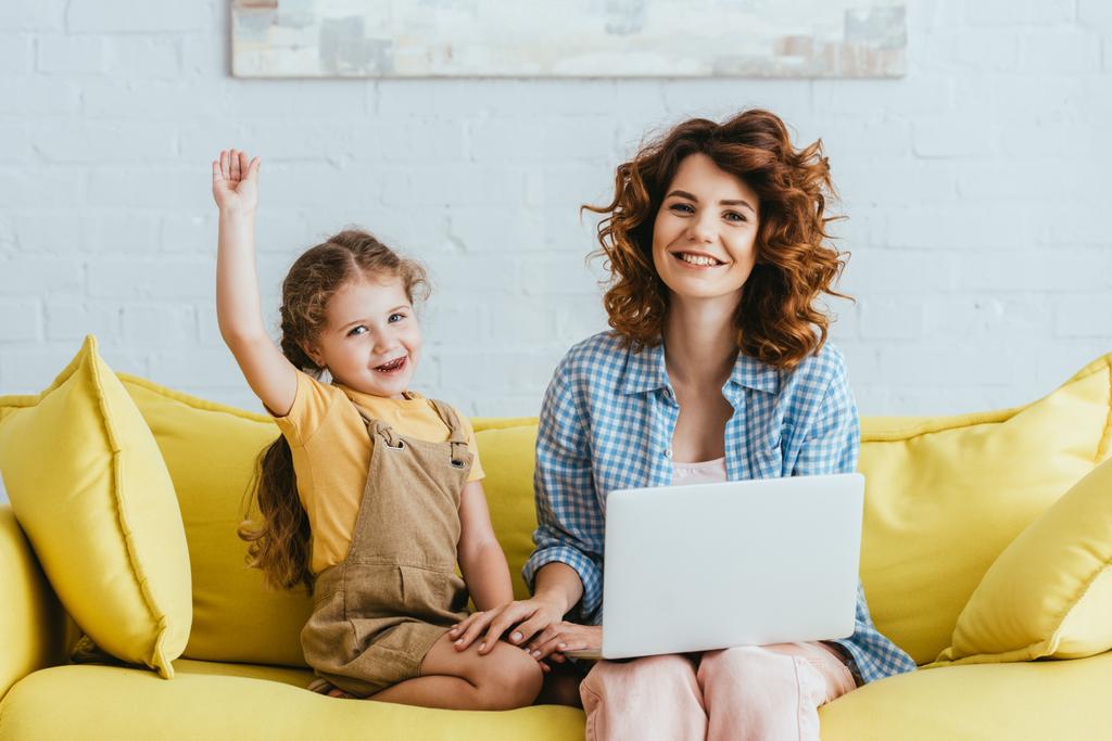 glückliches Kind, winkende Hand und junges Kindermädchen, das mit Laptop auf dem Sofa sitzt und in die Kamera lächelt - Foto, Bild