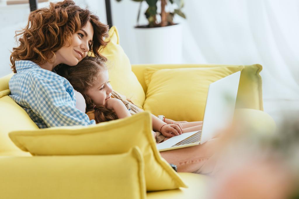 selektywne skupienie się na uśmiechniętej opiekunce i dziecku siedzącym na kanapie i korzystającym z laptopa - Zdjęcie, obraz