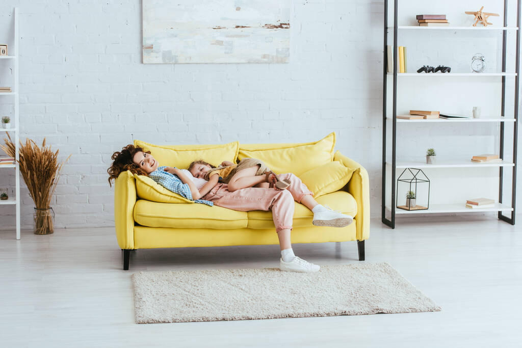 家でソファに横になりながらカメラで微笑む幸せな乳母と愛らしい子供 - 写真・画像