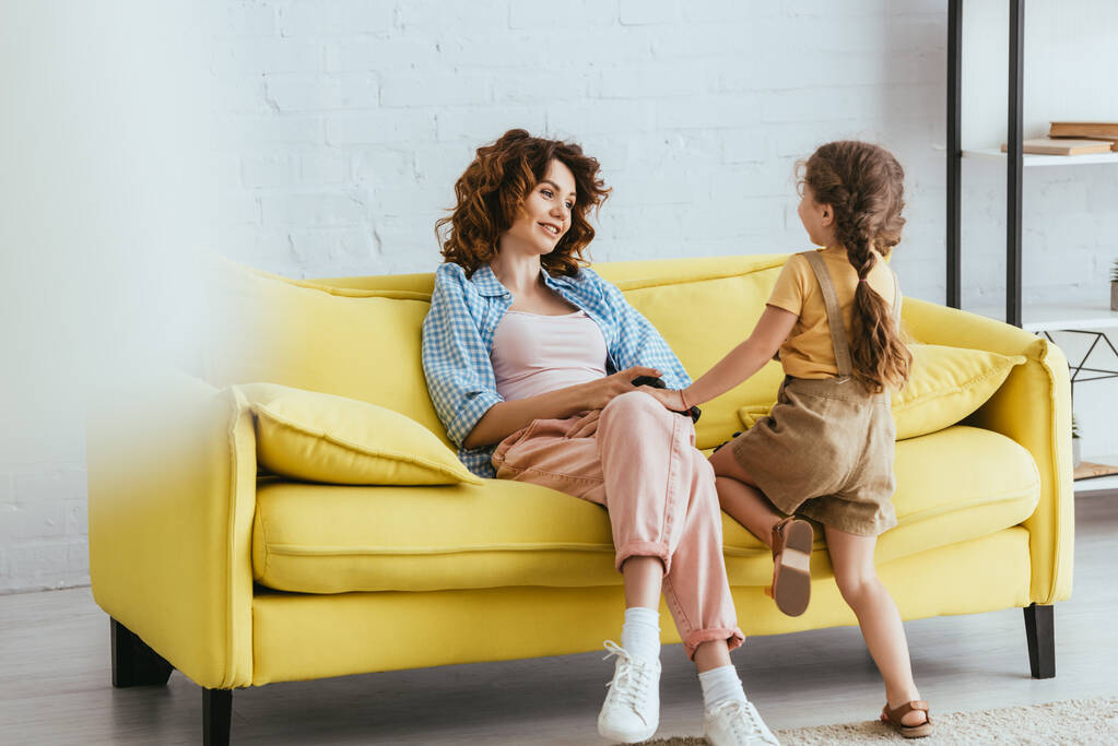 enfoque selectivo de la niñera sonriente cogida de la mano con el niño mientras está sentado en un sofá amarillo - Foto, Imagen
