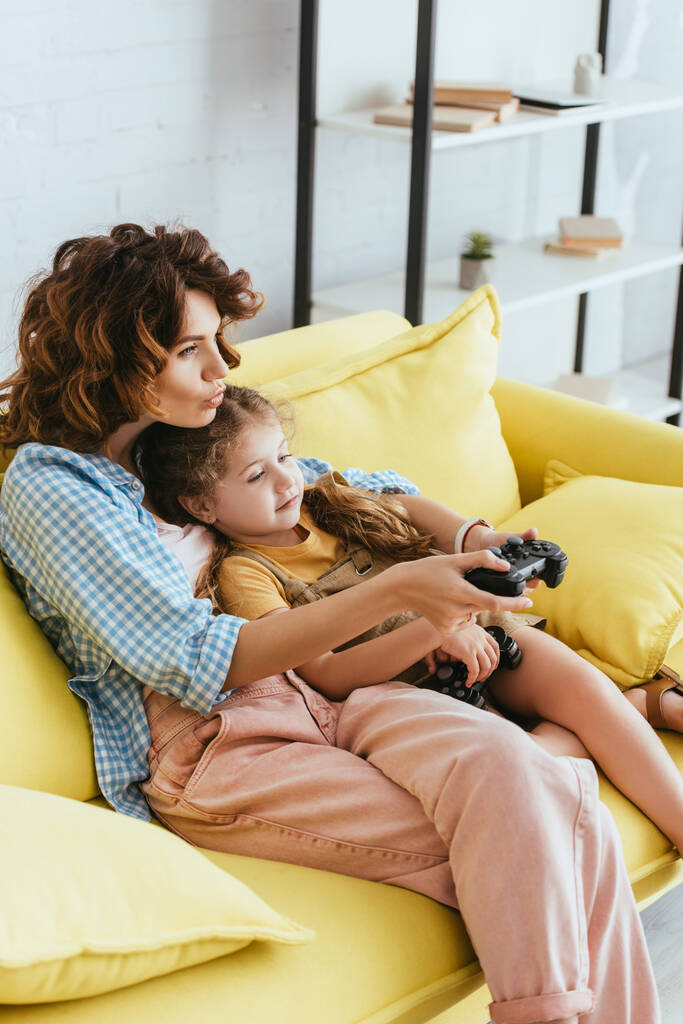 niñera y niño concentrados jugando videojuegos mientras están sentados en un sofá amarillo - Foto, imagen