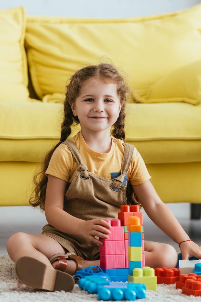 happy kid smiling at camera while sitting on floor near multicolored building blocks - Photo, Image