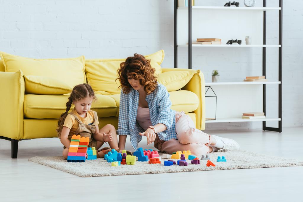 niñera joven y adorable niño jugando con bloques de construcción multicolores en el suelo cerca de sofá amarillo - Foto, imagen