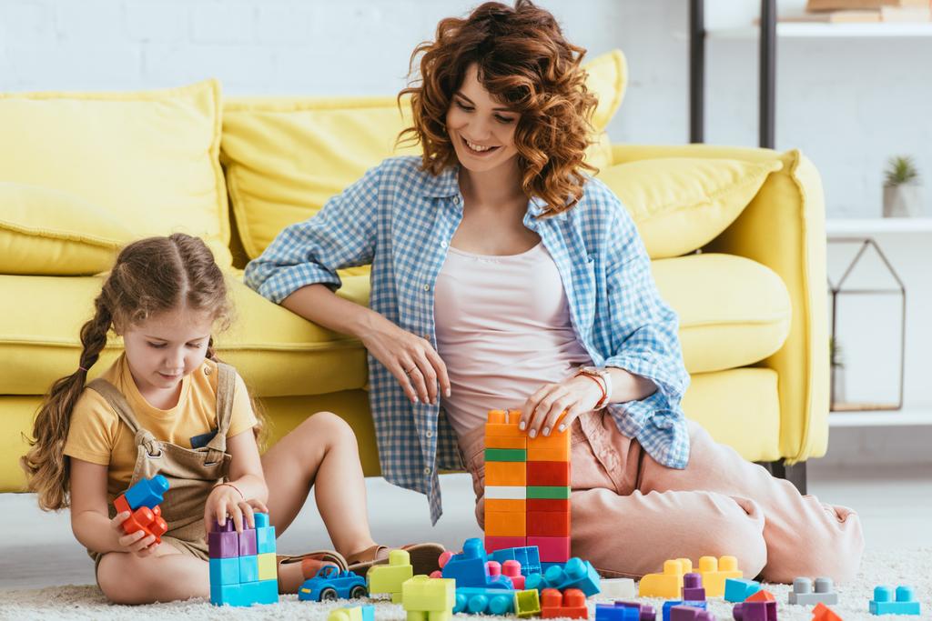 lachende nanny en schattig kind spelen met veelkleurige bouwstenen op de vloer - Foto, afbeelding