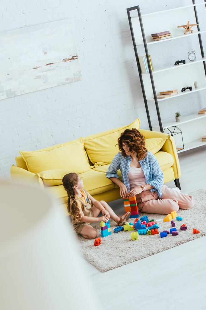selektiver Fokus von Kindermädchen und entzückendem Kind, das mit bunten Bausteinen auf dem Boden im Wohnzimmer spielt, Blick in den hohen Winkel - Foto, Bild