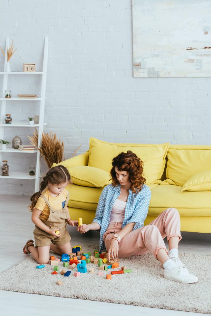 mladá chůva dávat blok k roztomilé dítě při hraní na podlaze v obývacím pokoji - Fotografie, Obrázek