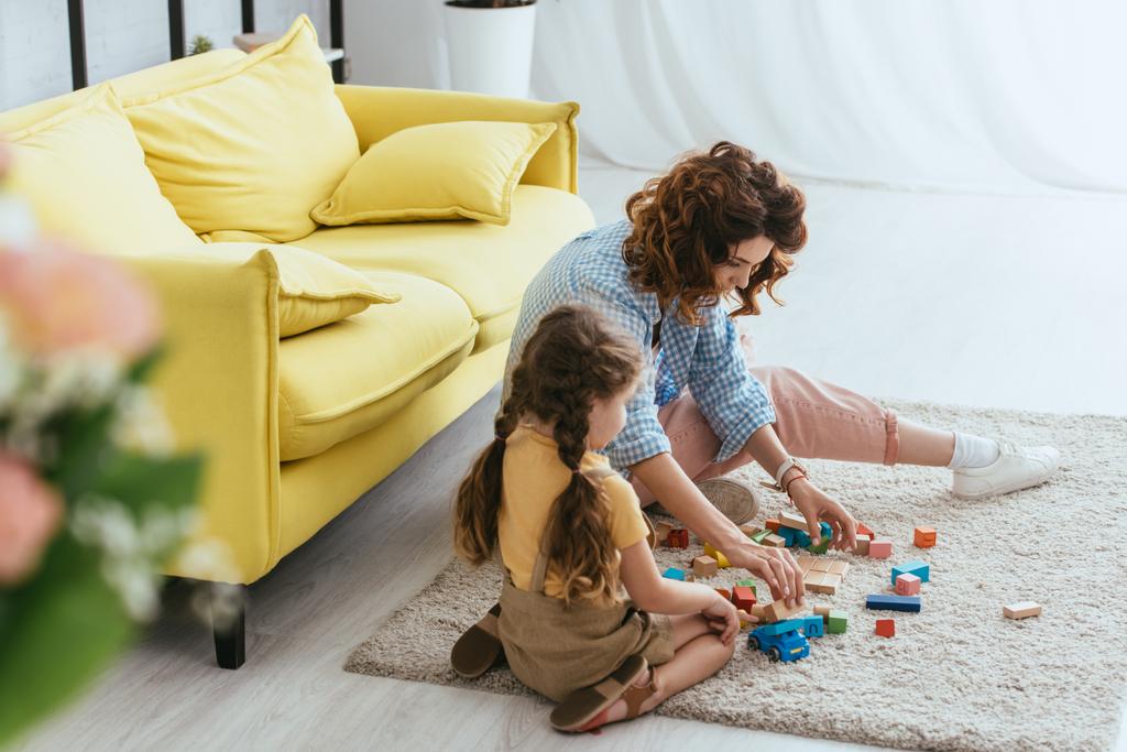 selektivní zaměření mladé chůvy a dítěte sedí na podlaze a hraje si s pestrobarevnými bloky - Fotografie, Obrázek