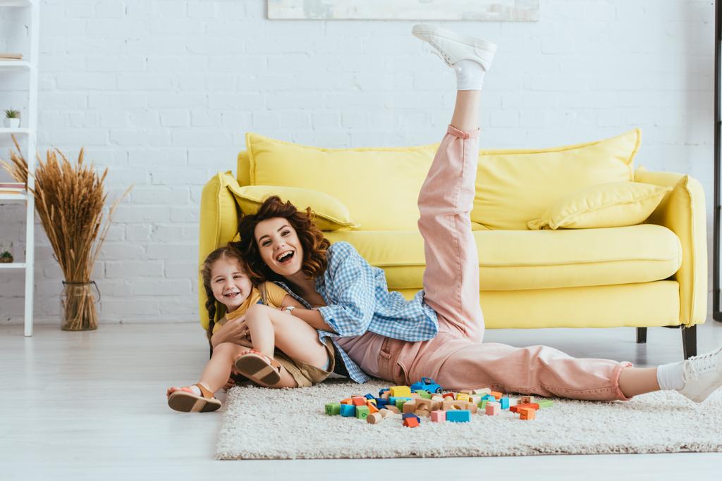 niñera alegre con la pierna levantada divertirse con el niño feliz en el suelo cerca de bloques multicolores - Foto, Imagen