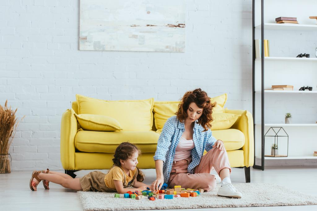 piękne niania i słodkie dziecko gra z wielobarwnych bloków na podłodze w pobliżu żółtej kanapie - Zdjęcie, obraz