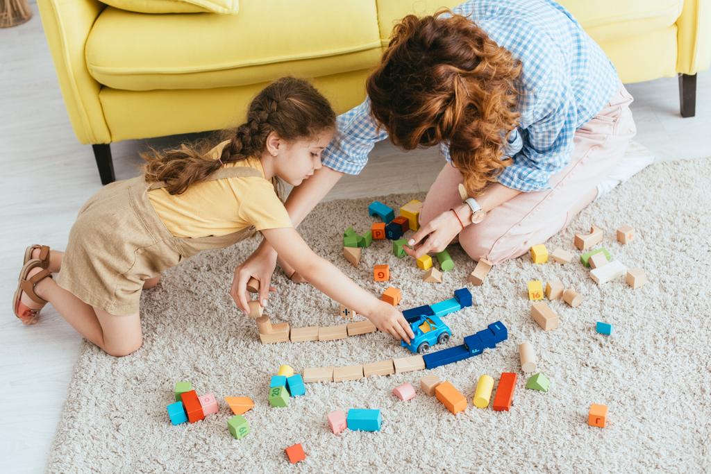 Bebek bakıcısının ve çocuğun çok renkli bloklar ve yerde oyuncak arabalarla oynadığı yüksek açılı manzara - Fotoğraf, Görsel
