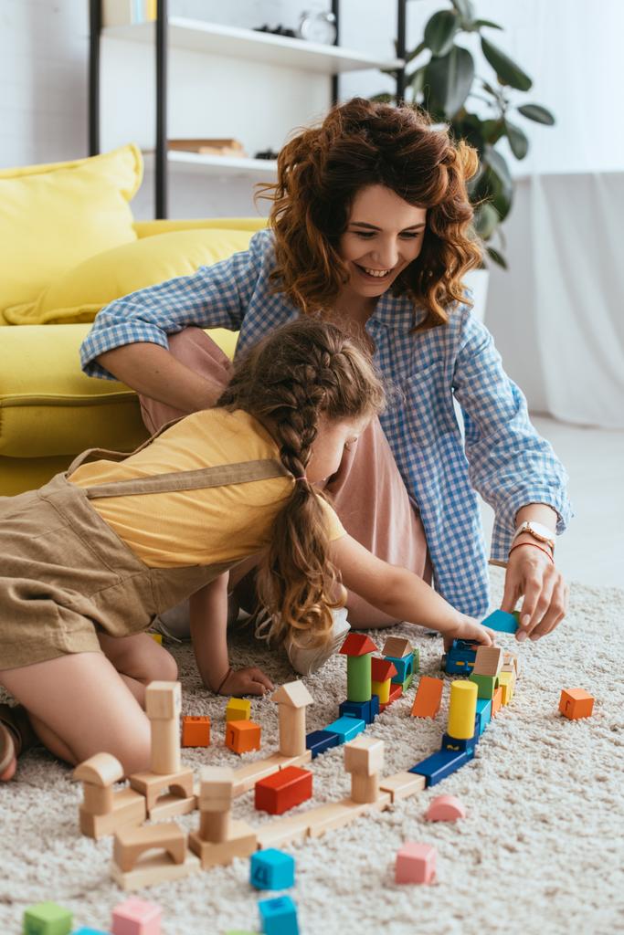 улыбающаяся няня с ребенком играет с разноцветными блоками, сидя на полу - Фото, изображение