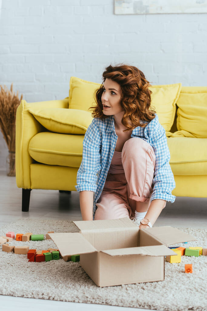 привлекательная молодая женщина сидит на полу возле коробки с разноцветными блоками - Фото, изображение