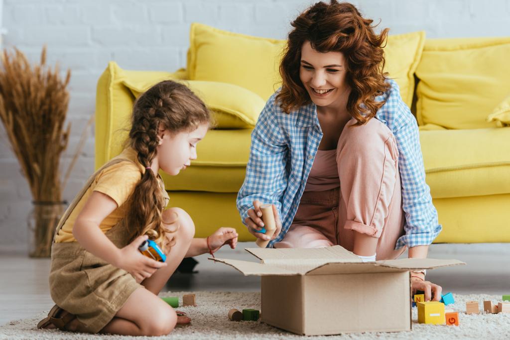 усміхнена няня і мила дитина сидить на підлозі біля картонної коробки з різнокольоровими блоками
  - Фото, зображення