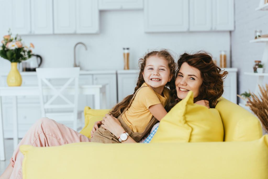 fröhliche Babysitterin und Kind lächeln in die Kamera, während sie sich auf dem Sofa in der Küche umarmen - Foto, Bild