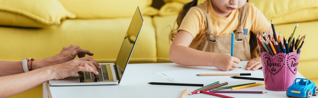 обрезанный вид детского рисунка с карандашом рядом с няней, работающей на ноутбуке, горизонтальное изображение - Фото, изображение