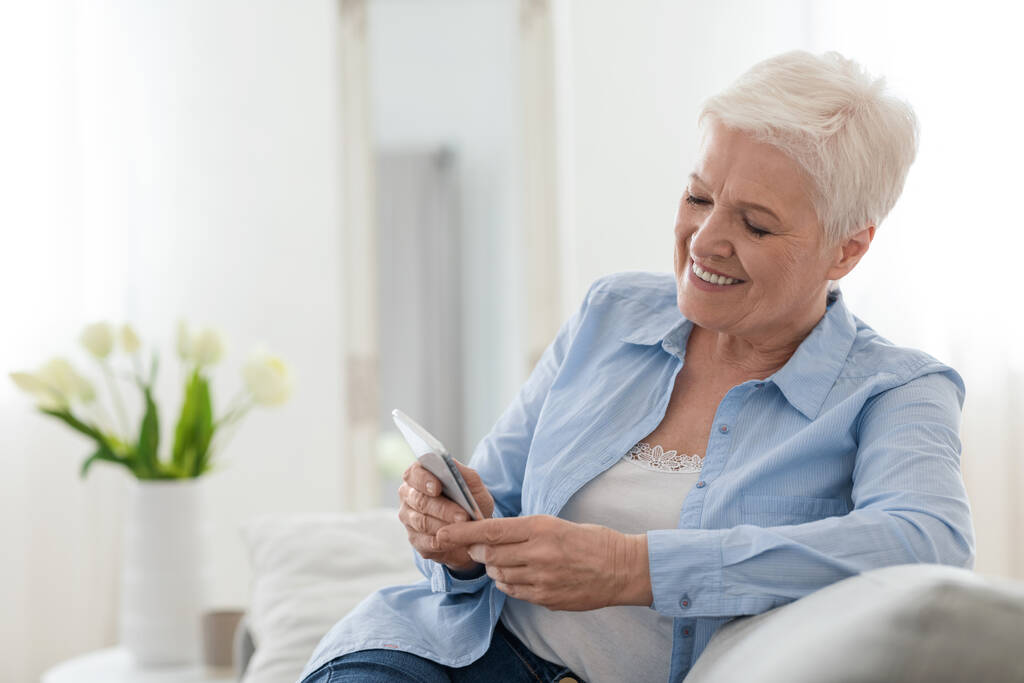 Happy elderly lady using mobile phone while relaxing on couch at home - Photo, Image