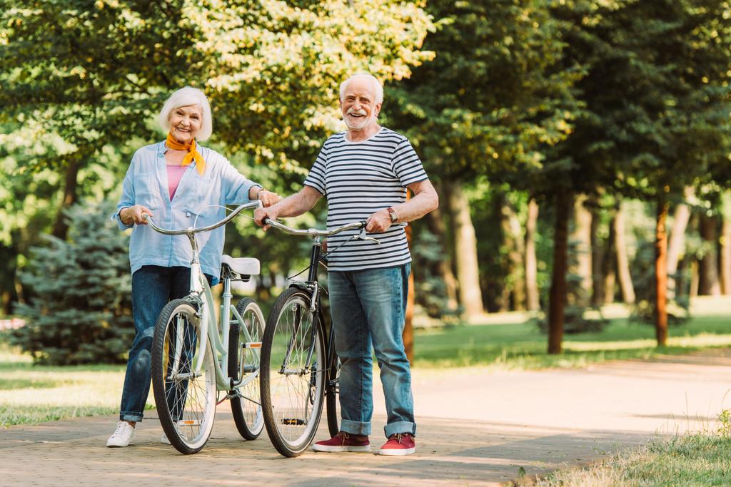 Пожилая пара улыбается в камеру во время прогулки на велосипедах по дорожке в парке  - Фото, изображение