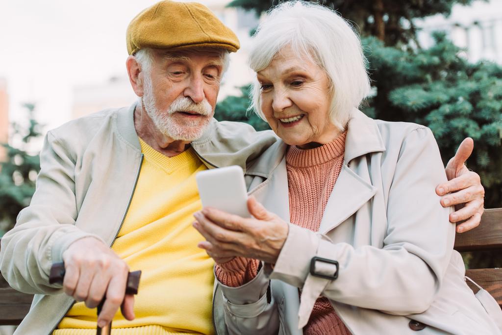 Enfoque selectivo de la mujer mayor sonriente sosteniendo teléfono inteligente cerca del marido en el banco en el parque
  - Foto, imagen