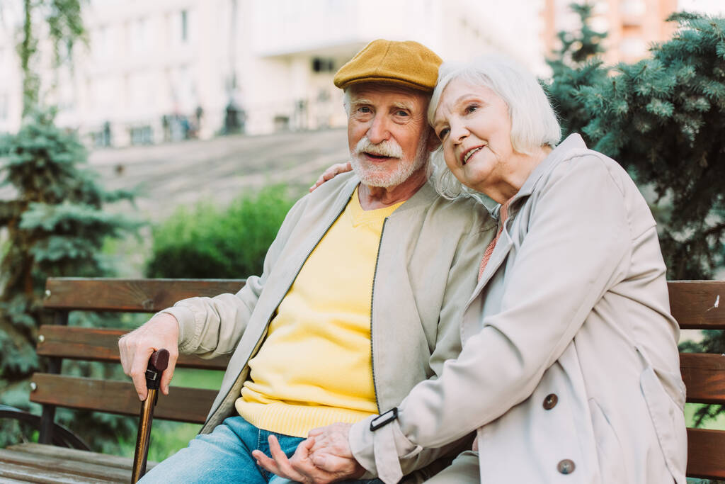 Enfoque selectivo del hombre mayor mirando la cámara mientras está sentado cerca de la esposa sonriente en el banco en el parque
  - Foto, Imagen