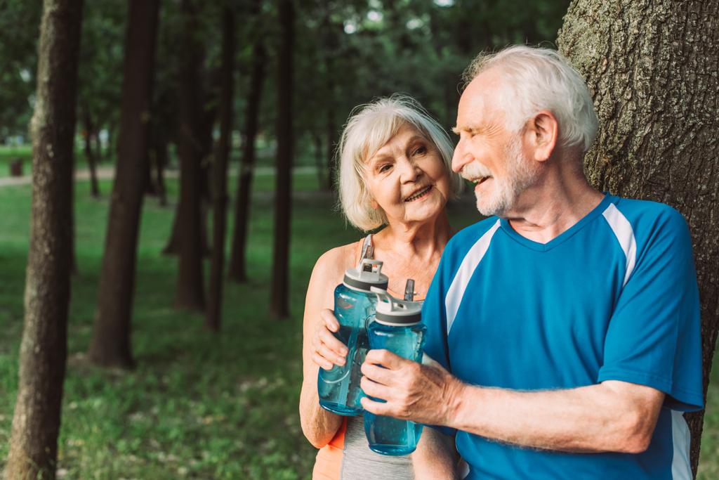 Couple âgé souriant tenant des bouteilles de sport dans le parc  - Photo, image