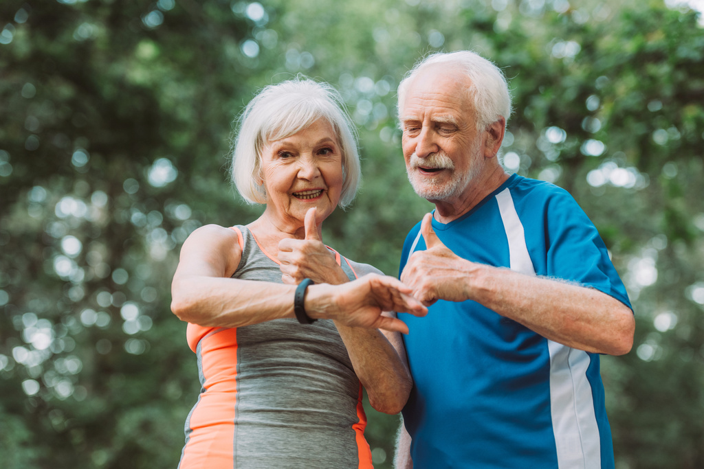 Usmívající se starší pár při pohledu na smartwatch a ukazující palce nahoru v parku  - Fotografie, Obrázek