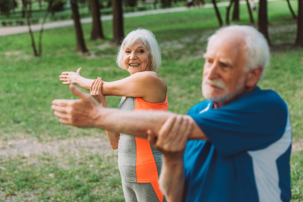 Επιλεκτική εστίαση της χαμογελαστή ηλικιωμένη γυναίκα άσκηση με τον σύζυγο στο πάρκο  - Φωτογραφία, εικόνα