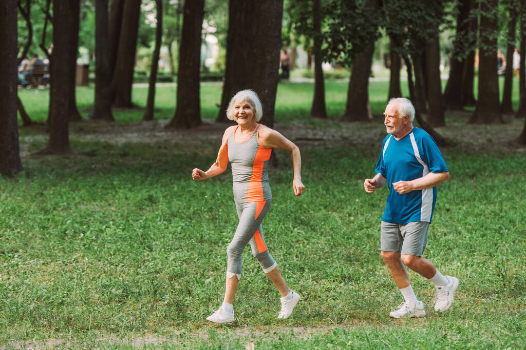 Sorridente donna anziana guardando la fotocamera mentre fa jogging vicino al marito nel parco  - Foto, immagini