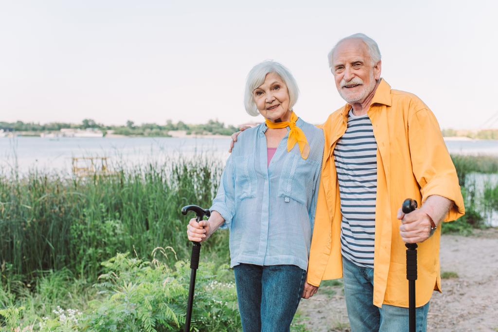 Sorridente uomo anziano abbracciando moglie con bastone da passeggio nel parco  - Foto, immagini