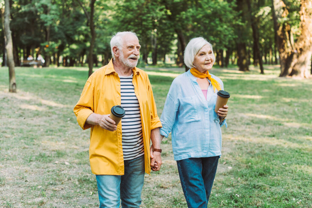 Улыбающаяся пожилая пара держит кофе, чтобы пойти во время прогулки в парке  - Фото, изображение