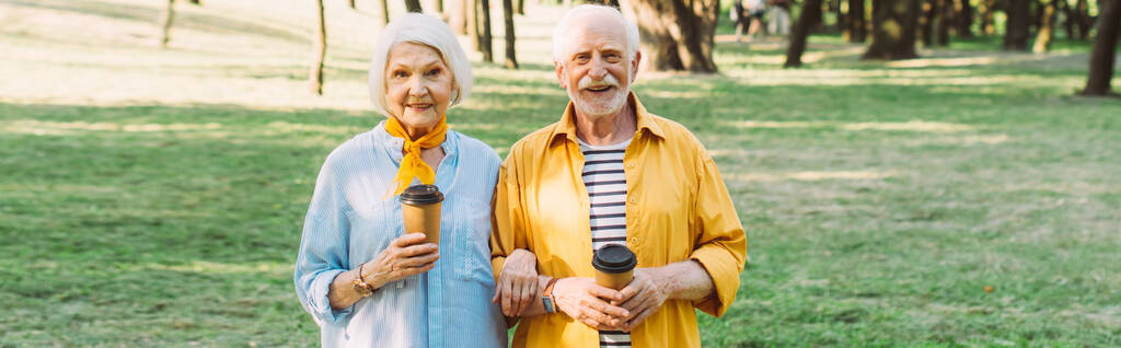 Recadrage horizontal de couples âgés souriants avec café pour aller regarder la caméra dans le parc  - Photo, image