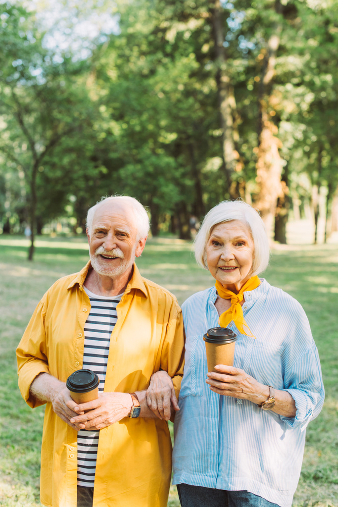 Usmívající se žena drží kávu jít a usmívá se na kameru poblíž staršího manžela v parku  - Fotografie, Obrázek
