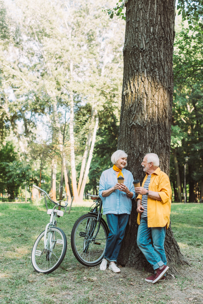 Couple âgé se souriant tout en tenant des tasses en papier près de l'arbre et des vélos dans le parc  - Photo, image