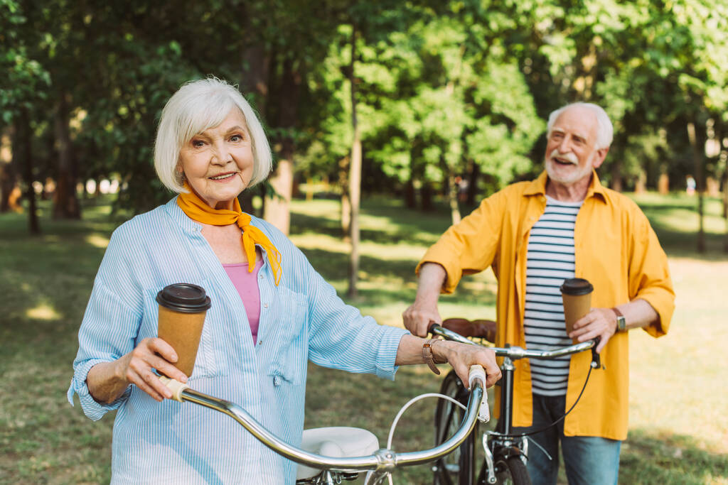 Επιλεκτική εστίαση της ηλικιωμένης γυναίκας κρατώντας χάρτινο κύπελλο κοντά ποδήλατο και ο σύζυγος στο πάρκο  - Φωτογραφία, εικόνα
