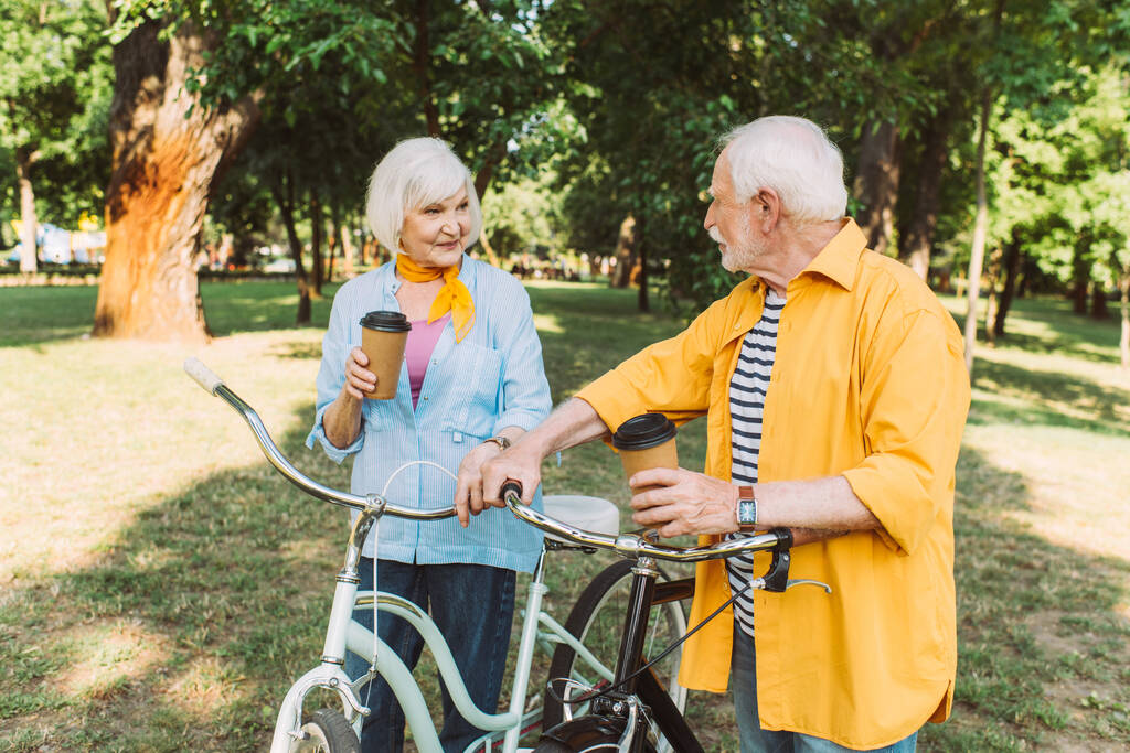 Couple aîné souriant avec café à emporter et vélos dans le parc  - Photo, image