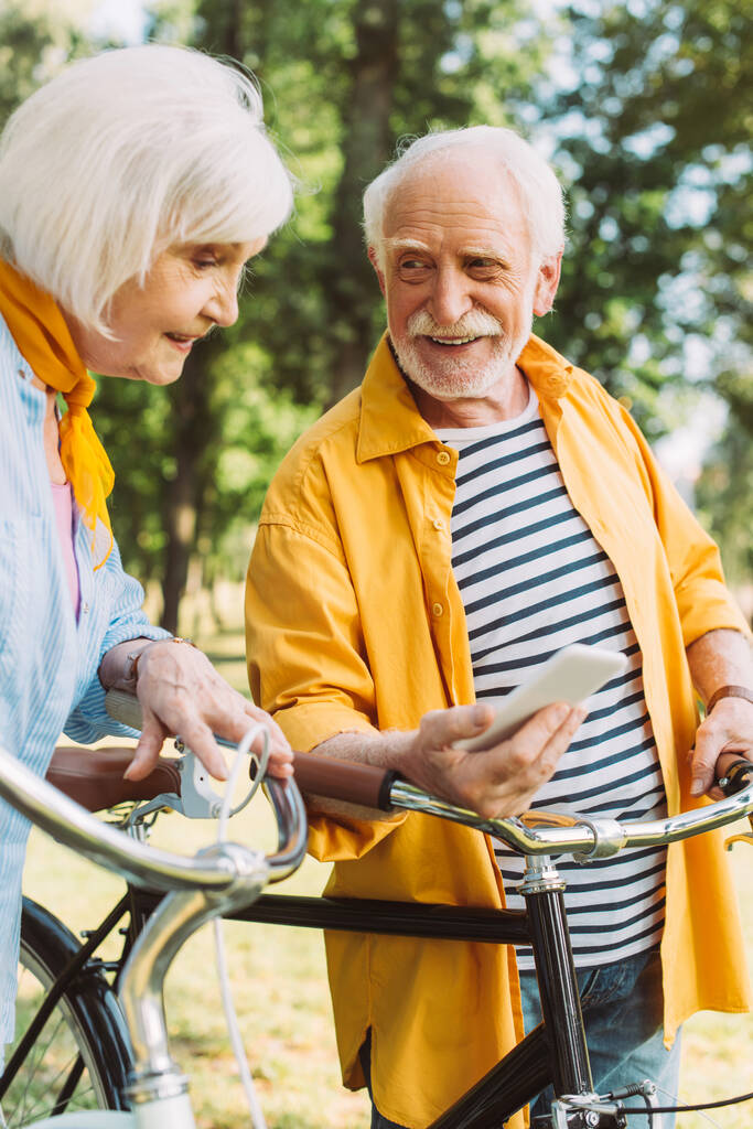 笑顔の妻と公園で自転車の近くにスマートフォンを保持する高齢者の選択的な焦点  - 写真・画像