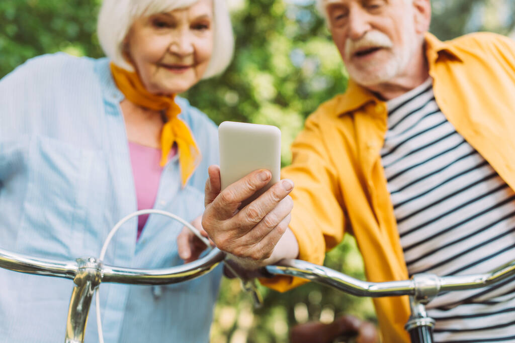 公園内の自転車の近くのスマートフォンを使用して高齢者のカップルの選択的な焦点  - 写真・画像