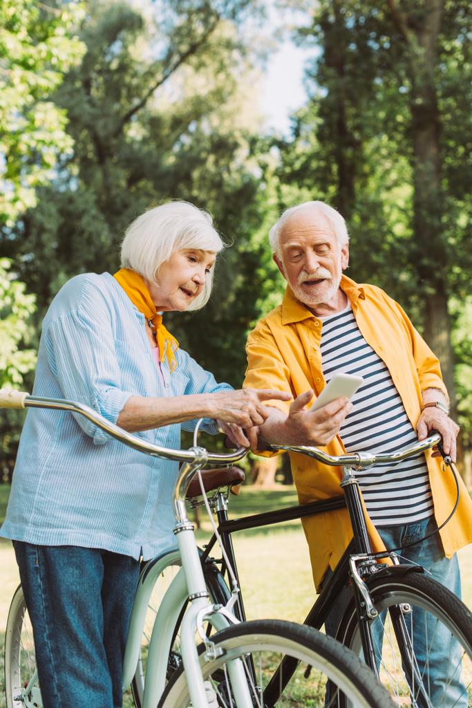 Starsza kobieta wskazująca na smartfona w pobliżu męża i rowery w parku  - Zdjęcie, obraz