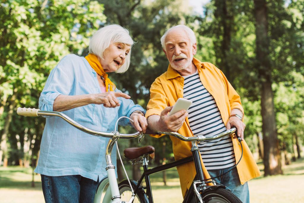 Oudere vrouw wijst naar smartphone naast man en fietsen in het zomerpark  - Foto, afbeelding
