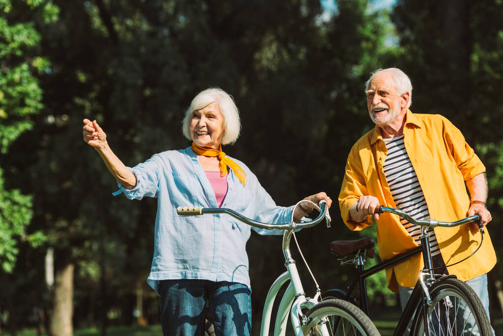 Ηλικιωμένη γυναίκα δείχνει με το δάχτυλο κοντά χαμογελαστός άνδρας και ποδήλατα στο πάρκο  - Φωτογραφία, εικόνα