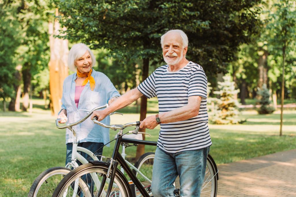 Улыбающийся мужчина смотрит в камеру во время прогулки с велосипедом рядом с женой в парке  - Фото, изображение