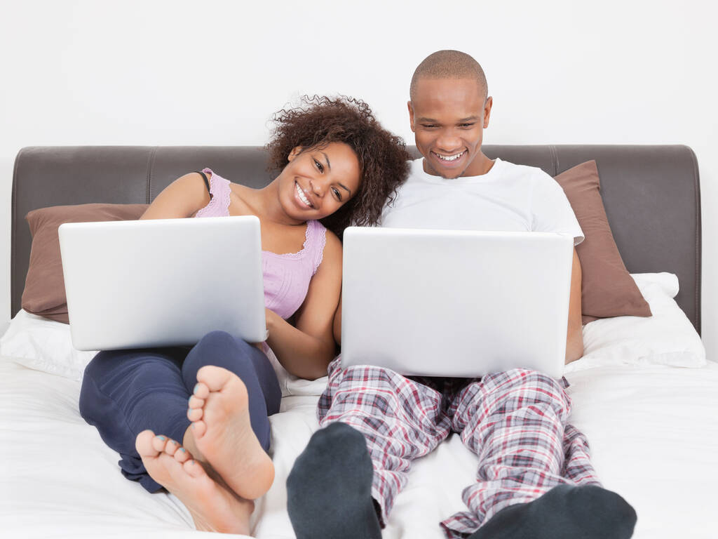 Yatakta dizüstü bilgisayar kullanan mutlu, genç bir Afrikalı çift. - Fotoğraf, Görsel