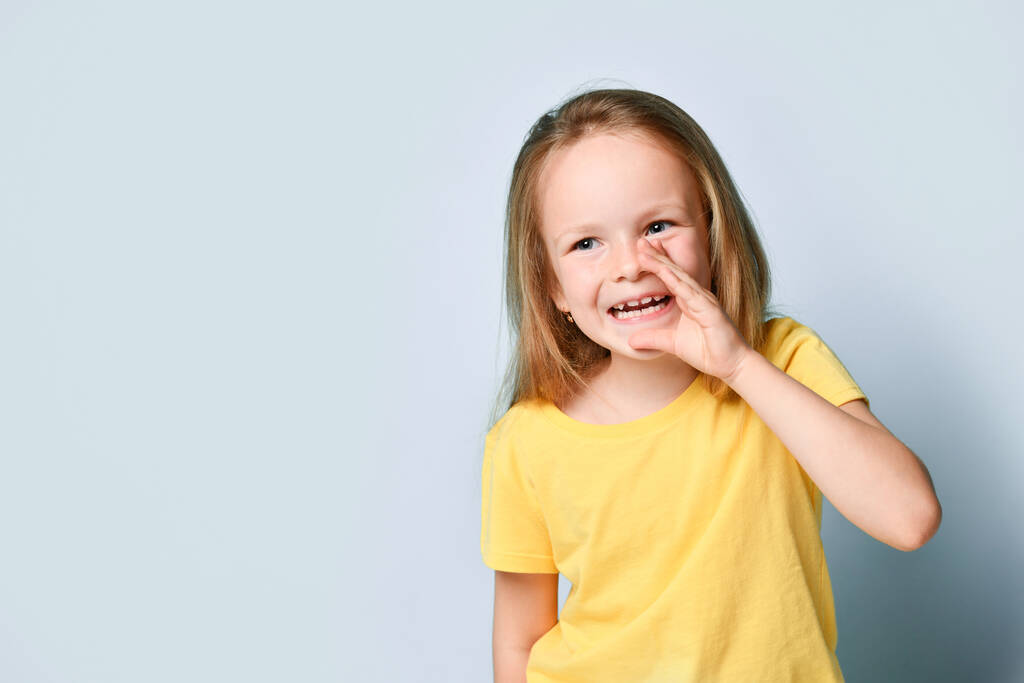 Милая маленькая девочка в ярко-желтой футболке, держащая руку близко к рту, делясь слухом или секретом. Талия до выстрела изолированы на светло-голубой, скопировать пространство - Фото, изображение